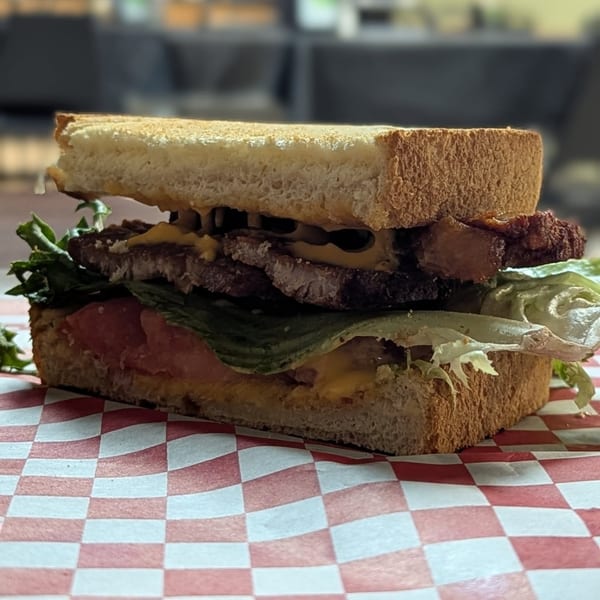 A thick sandwich with steak, lettuce, tomato, and cheese on toasted bread, on a red and white checkered paper.