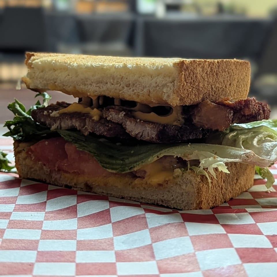 A thick sandwich with steak, lettuce, tomato, and cheese on toasted bread, on a red and white checkered paper.